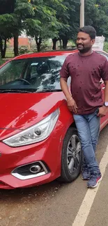 A man standing next to a red car on a tree-lined road.