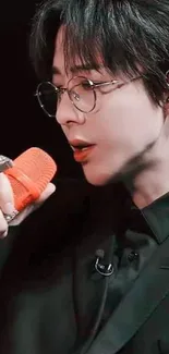 Singer with glasses holding an orange microphone in black outfit.