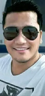 Person wearing sunglasses smiles in outdoor selfie with modern backdrop.