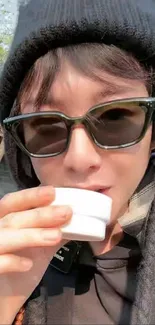 Person in sunglasses and beanie sipping from a cup outdoors.