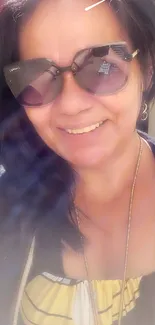 A woman in sunglasses smiles brightly in the sunlight, creating a summer selfie moment.