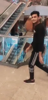 Man in black outfit walking in a mall with escalators and glass decor.