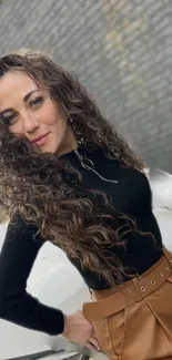 Woman with curly hair and black top poses stylishly against a brick wall.