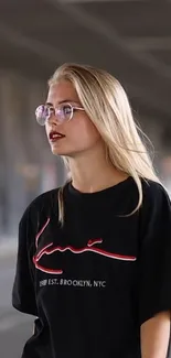 Blonde woman in sunglasses and a black graphic tee in an urban setting.