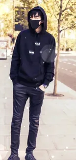 Man in black hoodie and mask standing on sidewalk.