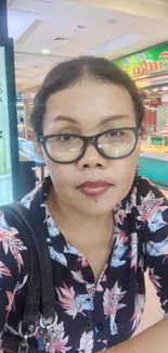 Woman wearing glasses at busy mall with floral shirt and vibrant background.