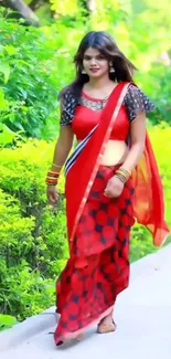 Woman in vibrant red saree walking amid lush greenery.