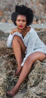 A stylish woman sitting on rocky beach scenery, wearing a white outfit.