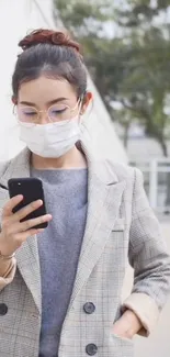 Woman wearing a mask and checkered blazer, using a smartphone outdoors.