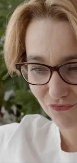 Woman wearing glasses and a white shirt, with green plants in the background.