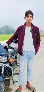 Young man in maroon jacket poses with motorcycle on a rural road.