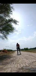 Person walks on sunlit countryside path under blue sky.