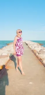 Woman walking on a pier towards the ocean under a clear blue sky.