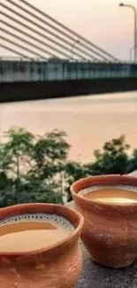 Two chai cups by a serene river and bridge at sunset.