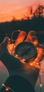 Hand holding a compass with glowing lights at sunset by a serene water backdrop.