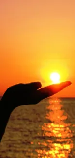 Hand holds the sun in a vibrant orange sunset over the ocean.