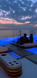 A couple relaxes by a pool under a colorful sunset sky.