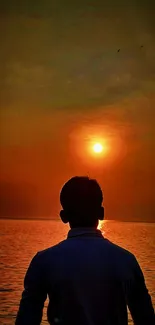 Silhouette at sunset over calm ocean with vibrant orange sky.