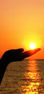 Hand silhouette holding sunset over ocean with orange sky