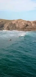 Surfers on teal ocean waves with rocky cliffs in the background.