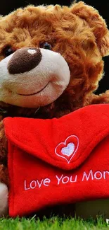 Cute teddy bear with red heart envelope in grass, reading 'Love You Mom!'