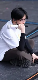 A person sitting thoughtfully on a dark surface, holding a white microphone.