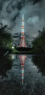 Tokyo Tower at night reflected in water, surrounded by greenery and clouds.