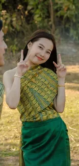 Cheerful woman in green traditional attire makes a peace sign in nature.