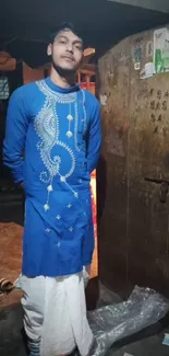 Man in blue embroidered kurta standing by a rustic door.