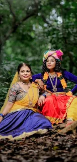 Two women in traditional attire sitting in a lush green forest.