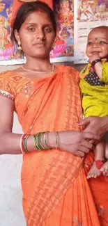 Indian woman in orange saree, holding a baby, with traditional posters in the background.