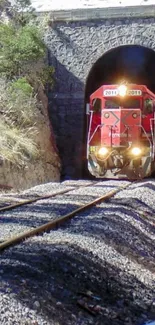 Red train exiting a stone tunnel into the sunlight.