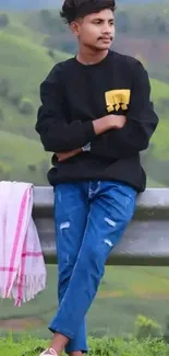 Young person standing by a scenic green hillside, wearing a black sweater and jeans.