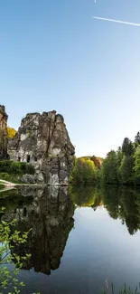 Tranquil lake reflecting towering rocks and greenery under a clear blue sky.