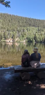 Two people sit by a peaceful lake with forest reflection.