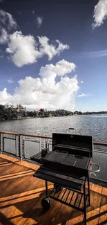 Mobile wallpaper of a BBQ grill on a wooden deck by a serene lake with a cloudy sky.