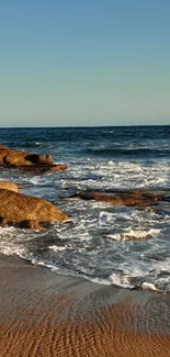 Serene ocean beach with waves and rocks under a clear blue sky.