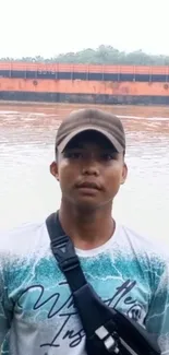 Man standing by a tranquil riverside, wearing a cap and casual attire.