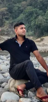 A man sitting peacefully on a rocky riverbank amidst nature.