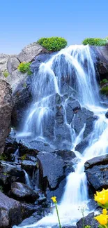 A serene waterfall in a rocky landscape with yellow flowers under a blue sky.