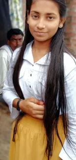 Stylish young woman in casual attire with long hair, posing against a brick wall.