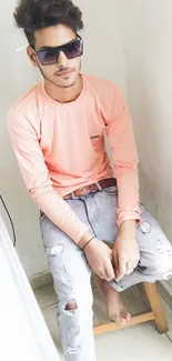 Young man in peach shirt and ripped jeans sitting casually on a chair.