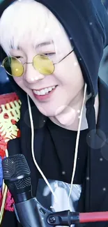 Person wearing a black hoodie and yellow glasses, smiling with a microphone.