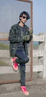 Trendy man in dark attire with bright sneakers by a fence.