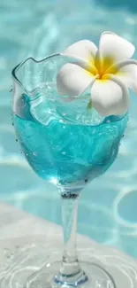 A clear glass with blue water and a white flower by the pool.