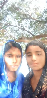 Two women stand under a large tree.