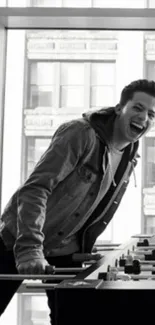 Black and white photo of a man playing foosball and laughing near large windows.