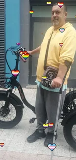 Man in yellow hoodie standing with electric bike against turquoise wall.