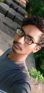 A young man in glasses takes a selfie with nature in the background.