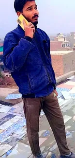 Man in blue jacket on rooftop with phone, vibrant cityscape background.